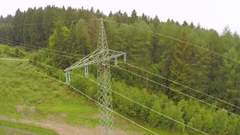 Un-Técnico-Trabajando-En-Una-Torre-De-Alto-Voltaje-En-Medio-De-Un-Exuberante-Bosque-Verde,-Vista-Aérea