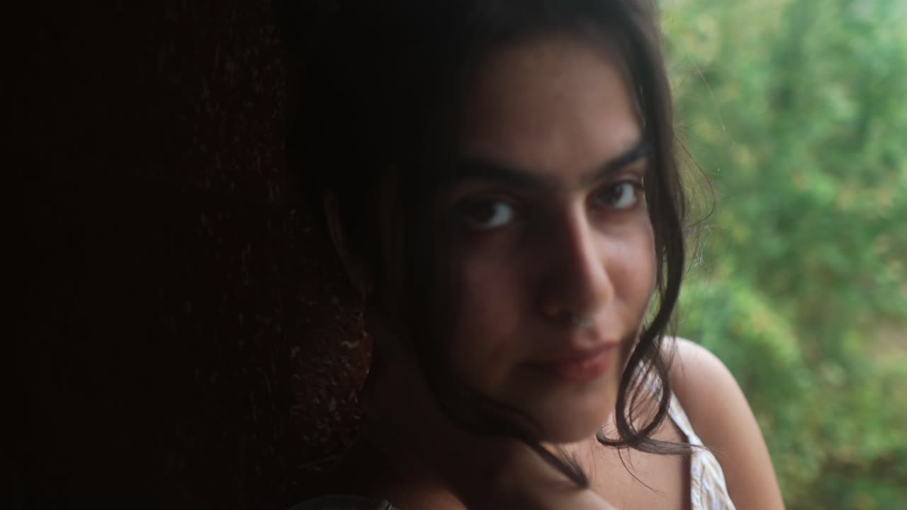 joven mirando sutilmente por encima de su hombro con una expresión serena, cerca de una ventana, vegetación borrosa en el fondo