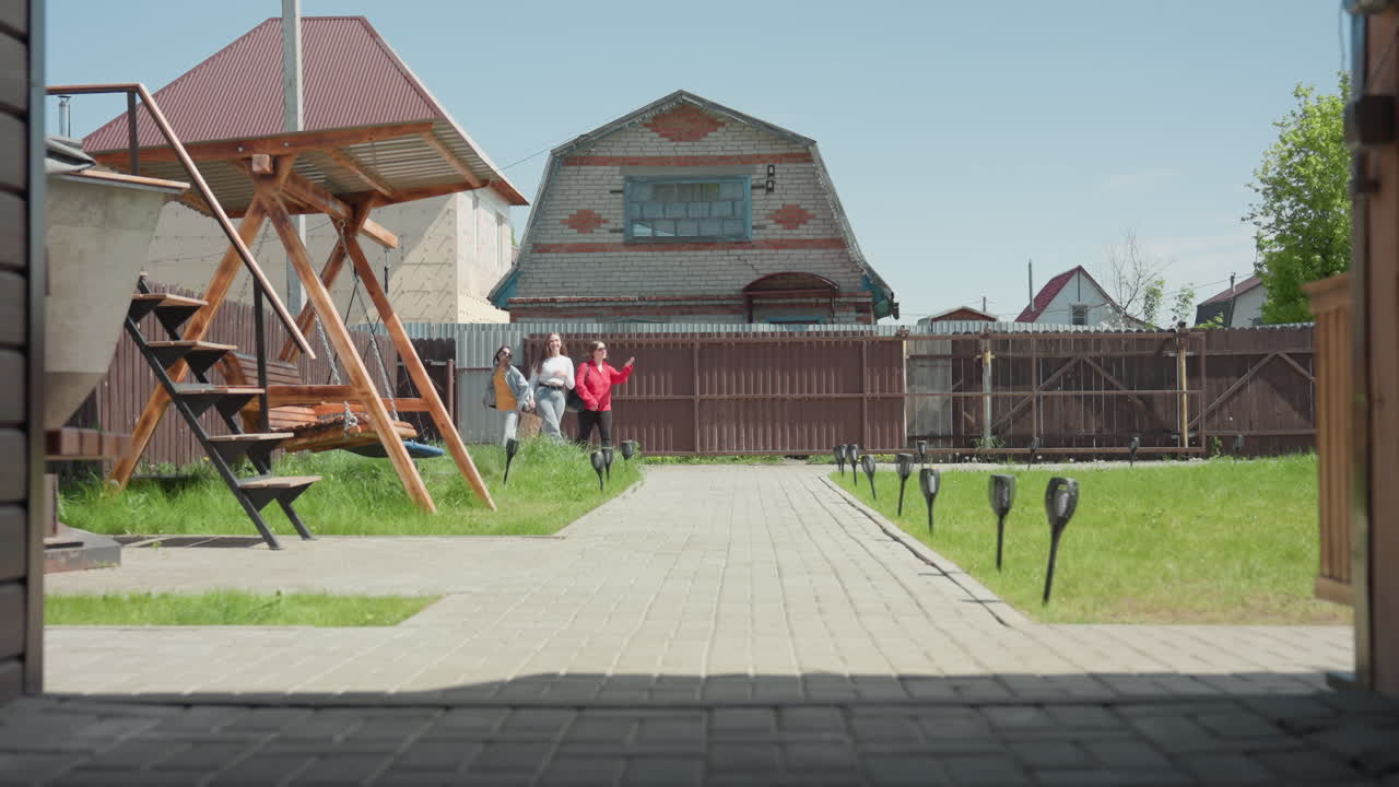 Young girls walk into recreational center along paved path, carrying bags and chatting, passing wooden swing and outdoor structure lined with small lights under bright sky in suburban yard