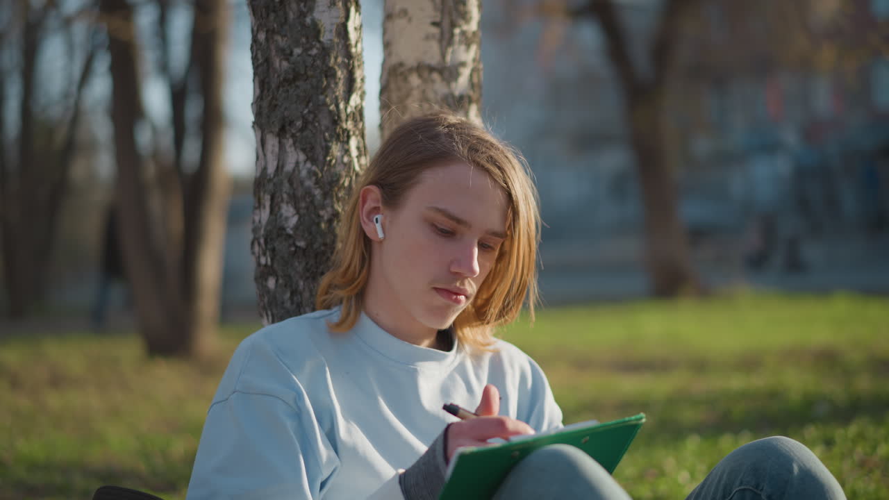 Tenåring organiserer studiemateriell ved siden av bjørk, ung student forbereder seg til timen i sterkt sollys, tenåring skriver og arrangerer klassenotater omhyggelig under skyggen av et bjørketre
