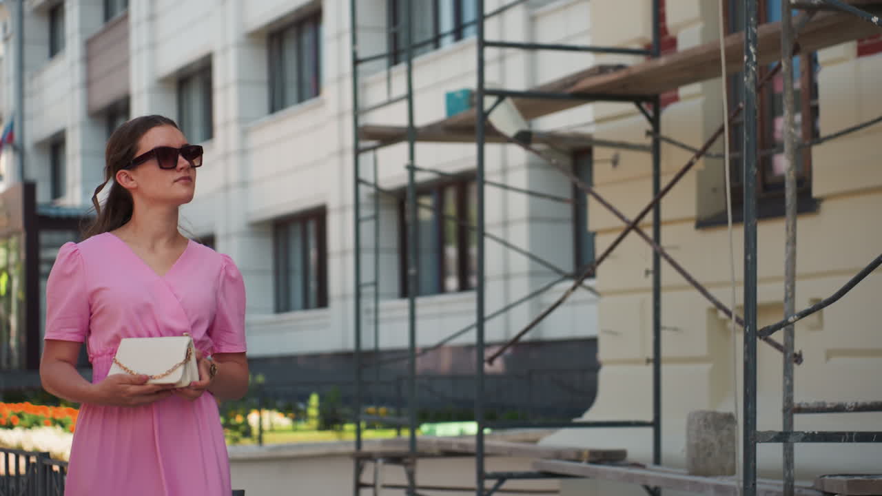 Mulher caucasiana com vestido rosa perto de andaimes, segurando uma clutch e usando óculos de sol, olhando para cima para a fachada do edifício, plano de fundo de renovação urbana, luz quente do meio-dia, expressão pensativa, moda