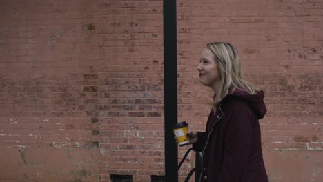 slow motion shot of a young carefree female walking down the street with a hot beverage in hand with a red brick wall in the background