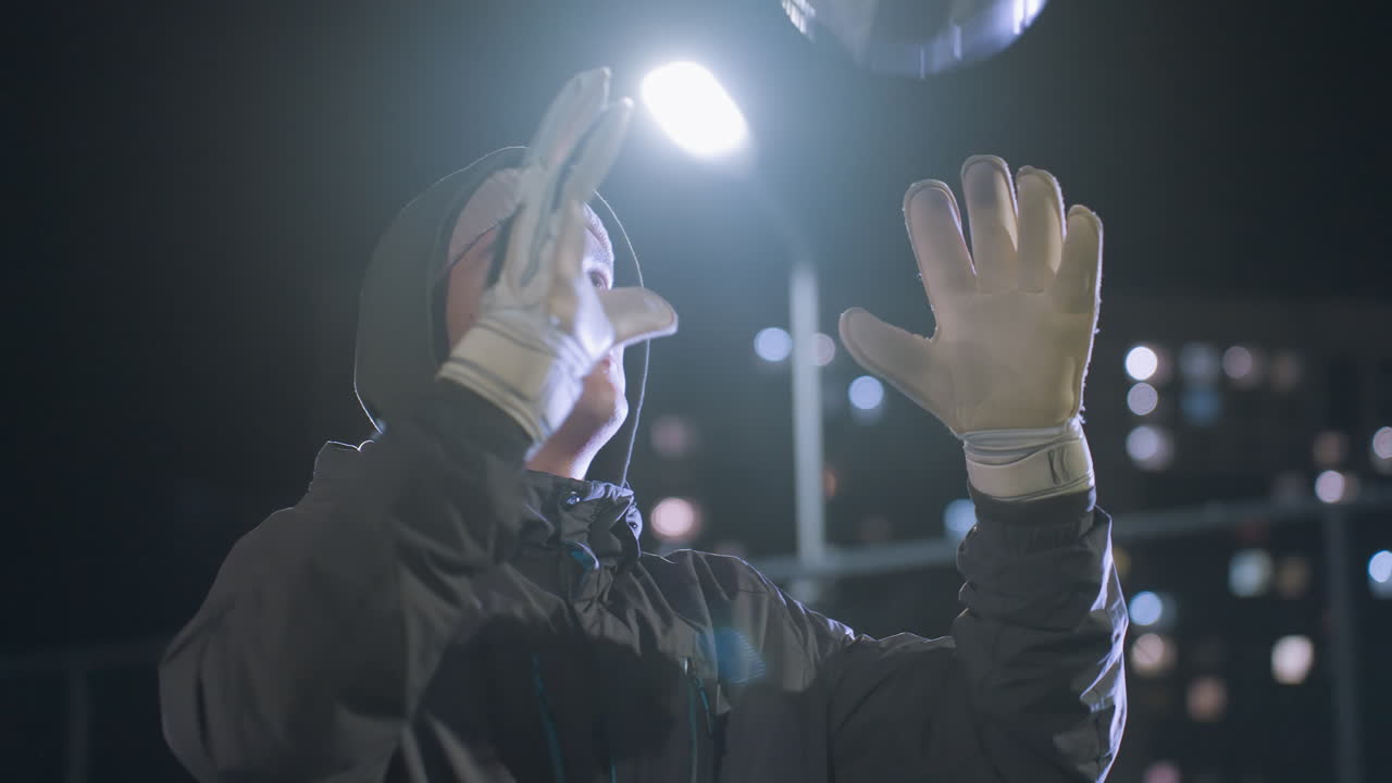 장갑을 입은 축구 선수가 공중으로 축구공을 던지고 그것을 잡는 클로즈업, 흐릿한 도시 배경, 저녁 불빛이 보케 효과를 만니다.