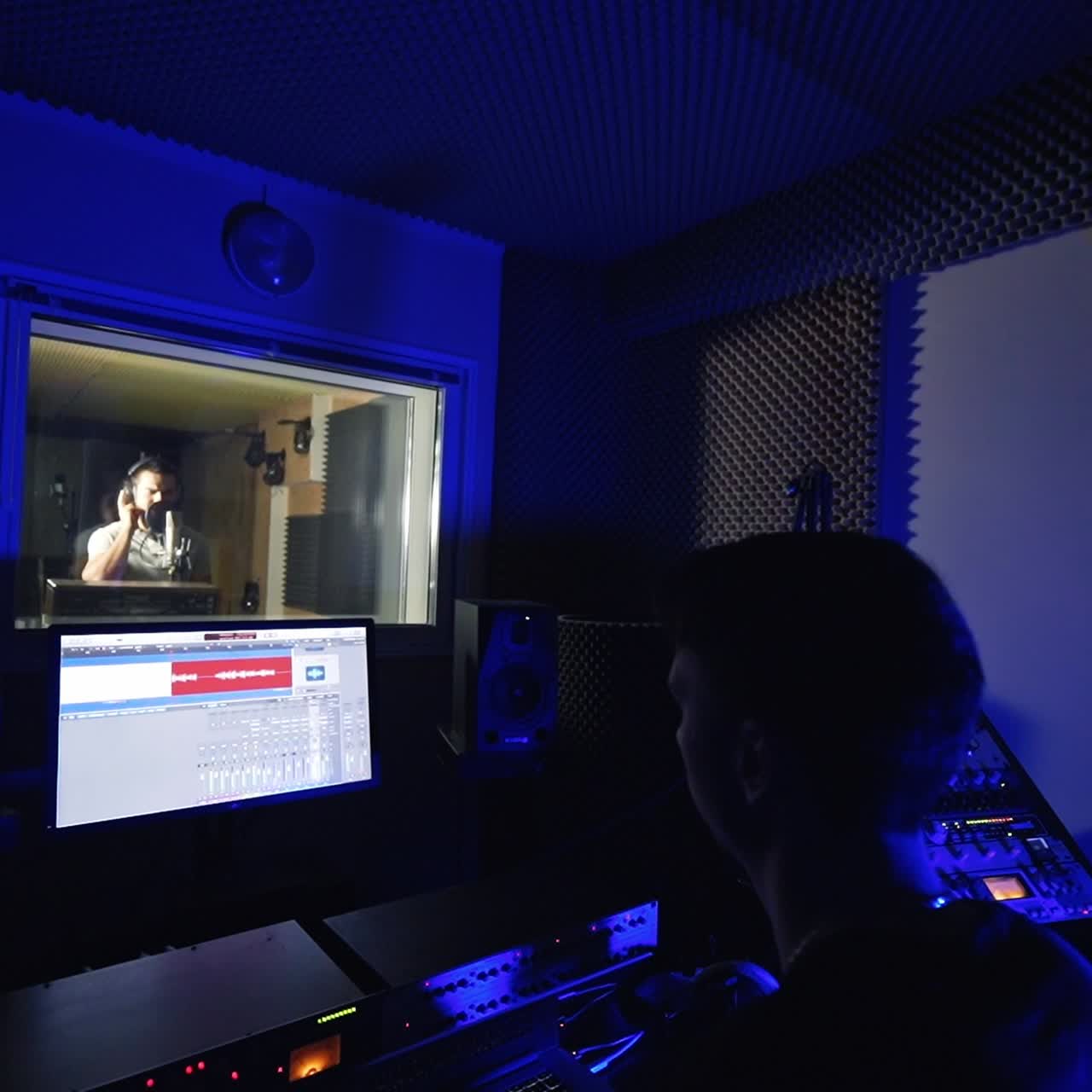 Modern professional studio for music recording. Sound engineer is sitting in front of equipment. Male singer performs a song in the next room behind a window