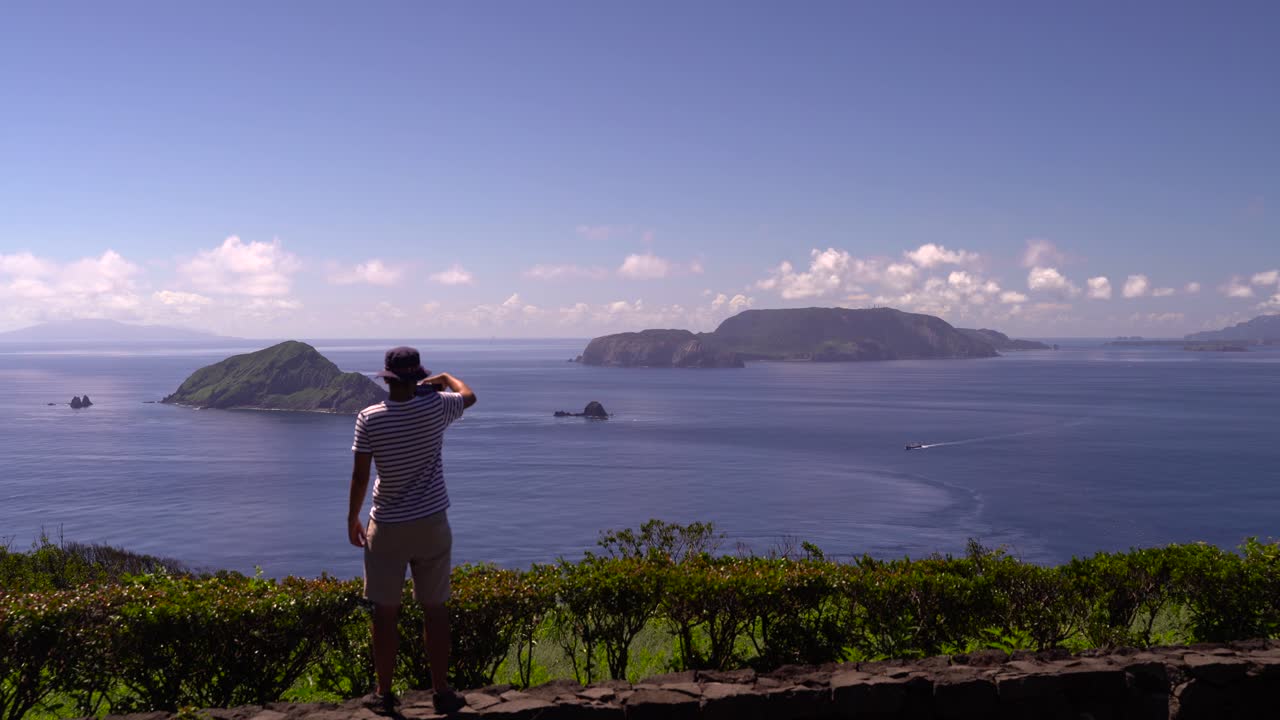 hombre que viaja solo tomando fotos con su teléfono inteligente mirando hacia el hermoso océano azul y las islas en un día claro y soleado