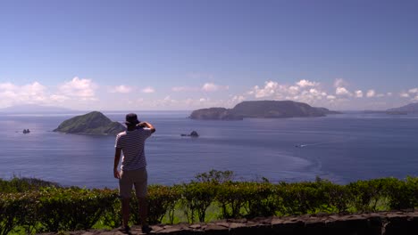 Male-solo-traveler-taking-pictures-with-his-smartphone-looking-out-towards-beautiful-blue-ocean-and-islands-on-clear-and-sunny-day