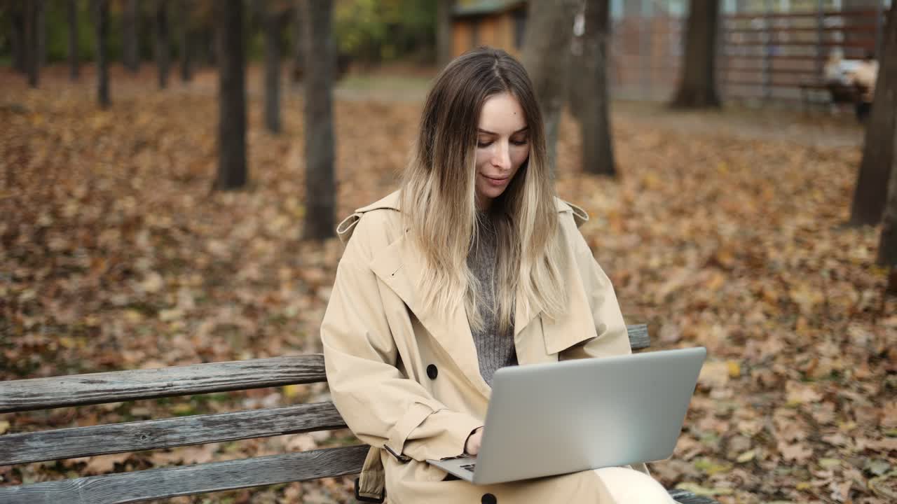 Young beautiful woman of European appearance works at a laptop, sitting on a park bench