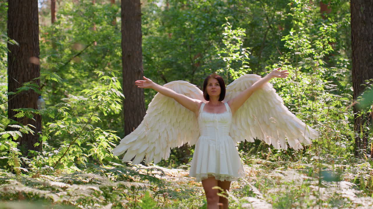 Fairy white lady with large feathered angel wings standing in lush green woodland lifting arms gracefully toward sunlight, surrounded by glowing leaves and soft forest light