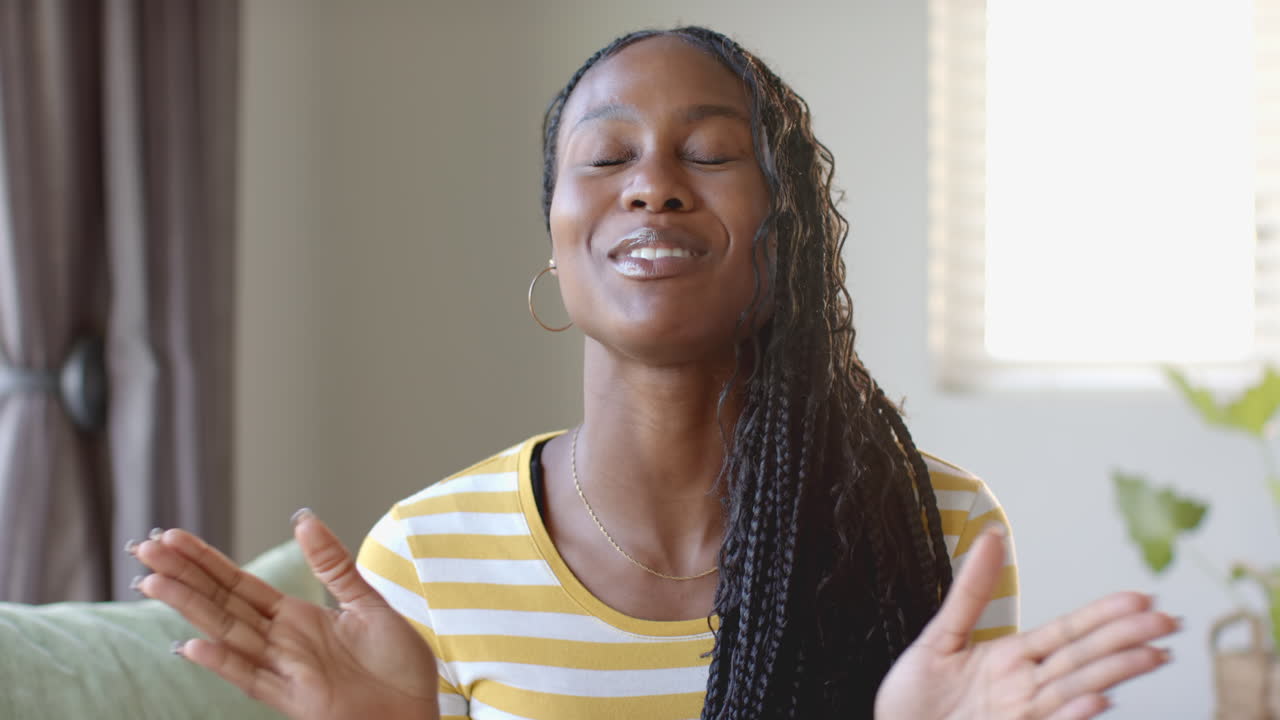 Smiling african american woman in striped shirt sitting on couch, talking on video call at home