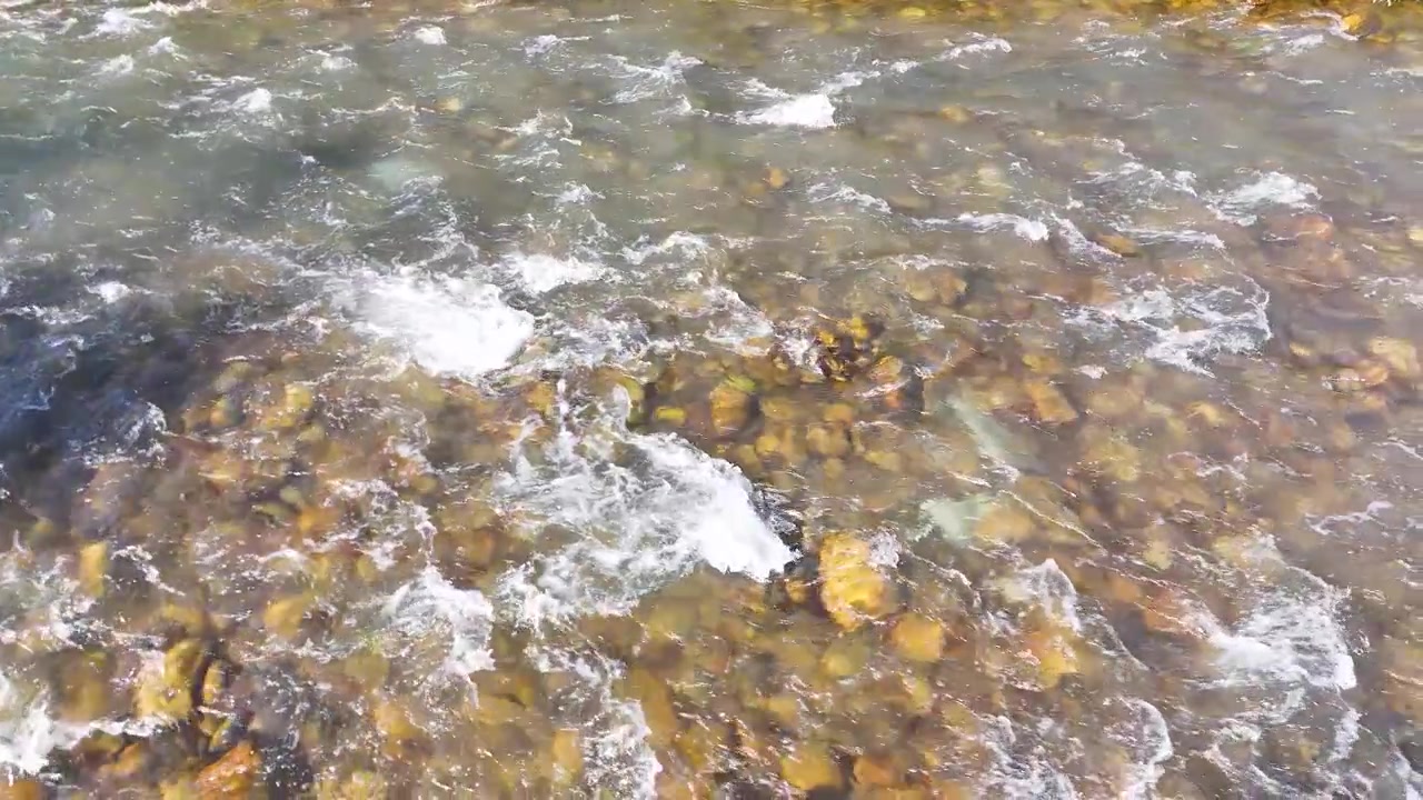 High-angle static shot of freshwater flowing rapidly over a rocky riverbed in bright daylight.