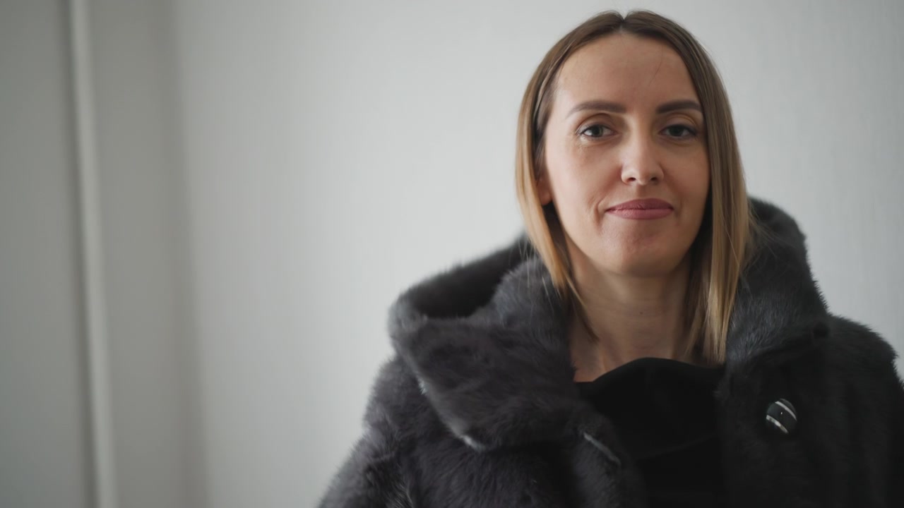 Nærbillede af kvinde med skulderlangt blondt hår iført sort off-shoulder top. Kvinden tager gradvist en mørkegrå pelsfrakke på og justerer den, hvilket afslører teksturdetaljer i begge hendes beklædningsgenstande