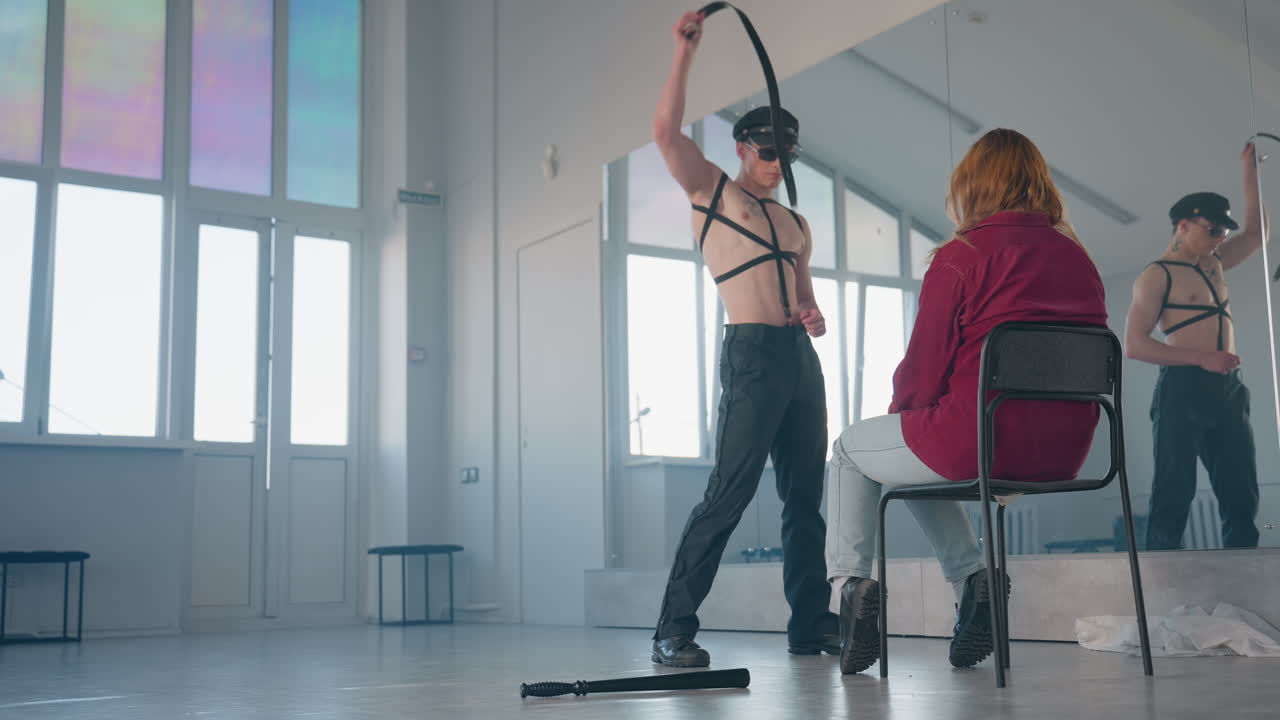 man removes belt then wraps strap around neck and tosses it aside in front of woman seated on chair in bright mirrored dance studio with baton and soft cloth on floor near reflection