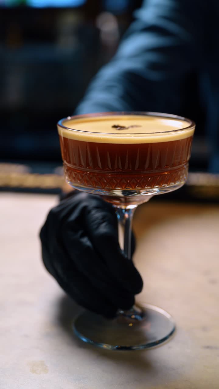 Barman con guanti neri che tiene lo stelo di un bicchiere da cocktail. La mano di un barman professionista che presenta un cocktail espresso martini sul bancone del bar.