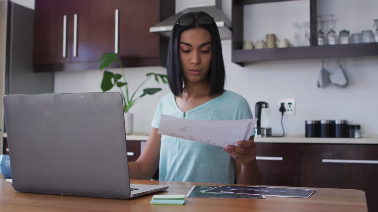 Persona gender fluid multietnica seduta alla scrivania, che prende appunti e lavora da casa usando un laptop.