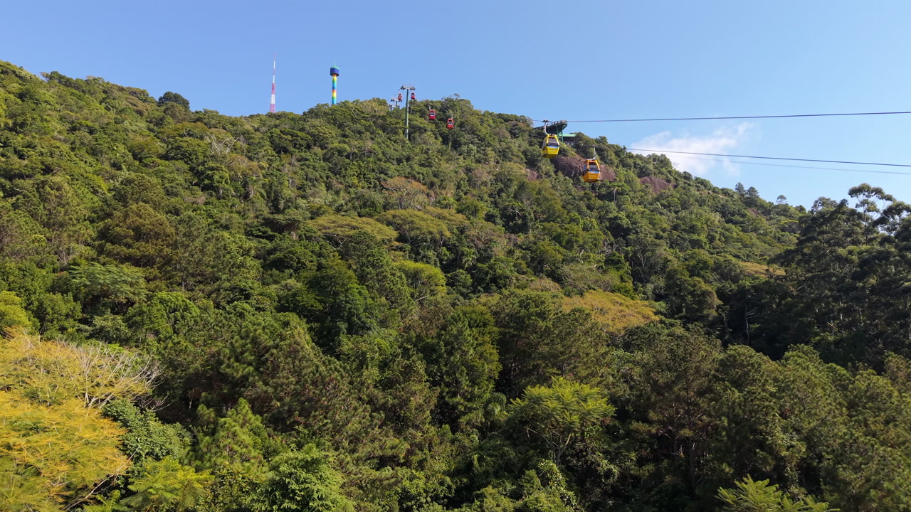 Flying alongside bright yellow cable car gondolas as they travel over a lush green forested mountain in Balneario Camboriu - Praia das Laranjeiras, Brazil on a sunny day