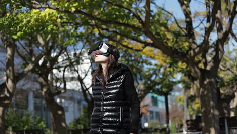 young dark-haired woman in warm black waistcoat wearing virtual reality glasses among green trees