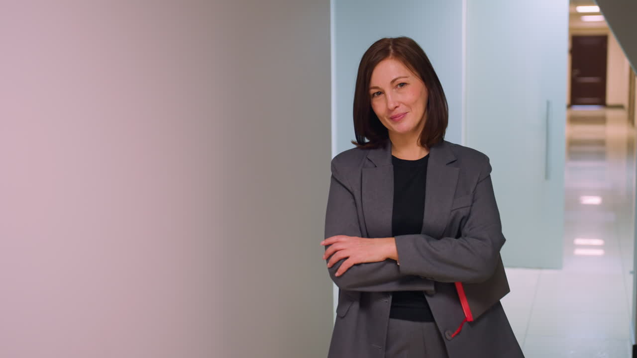 Giovane donna in abbigliamento professionale che tiene una cartellina in mano, gesticola con sicurezza e un sorriso rilassato, mentre cammina lungo il corridoio di un ufficio moderno, professionale e sicura di sé