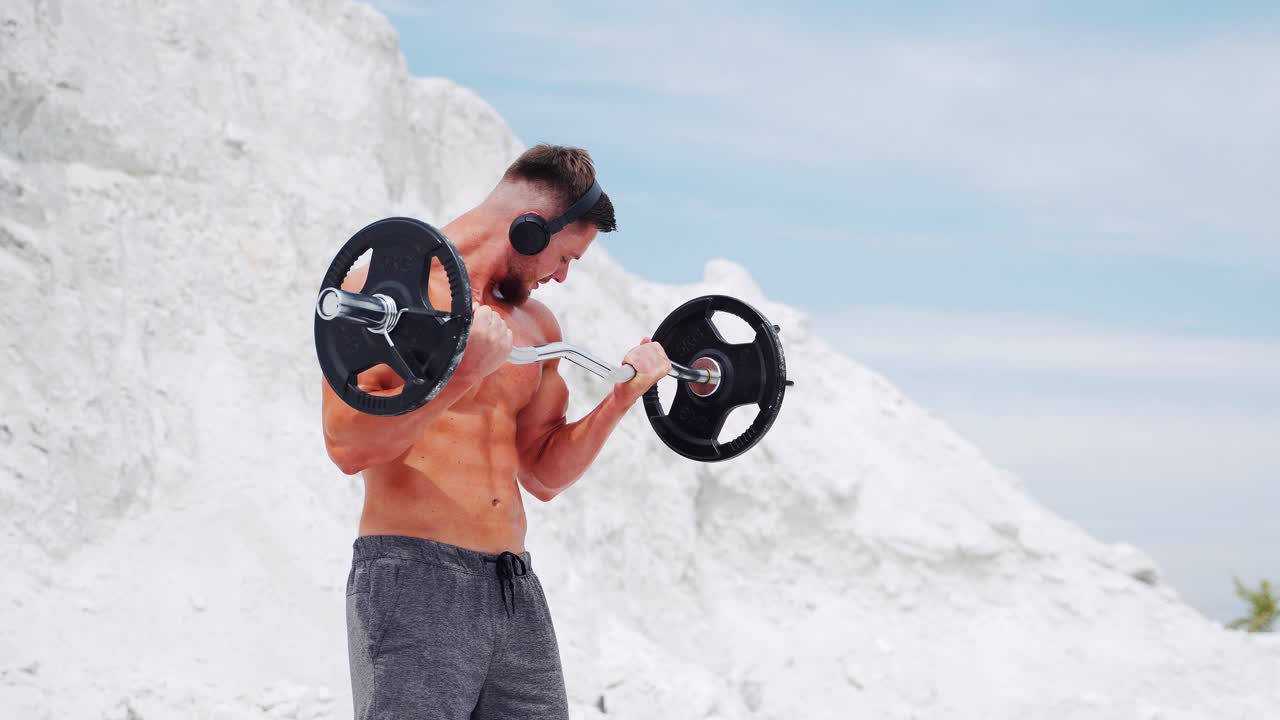 Shirtless man in headphones trains barbell for biceps. Outdoor sports