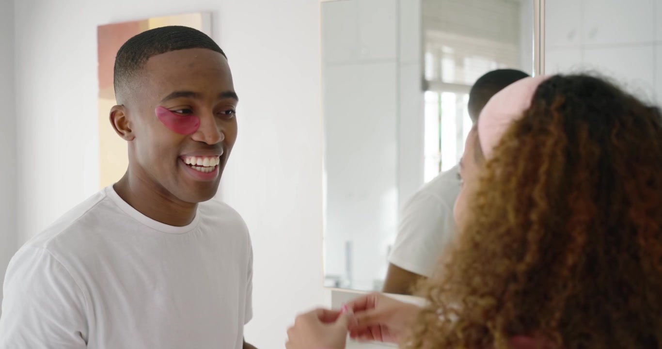 Couple at mirror, woman reaching, wearing headband applying pink eye patches for morning care