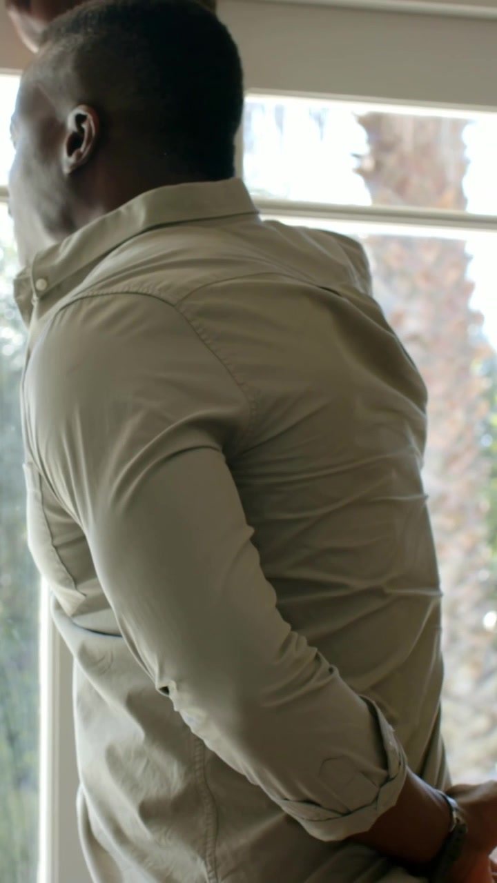 Vertical video: Touching blinds, man clasping hands behind at window in khaki shirt watching trees