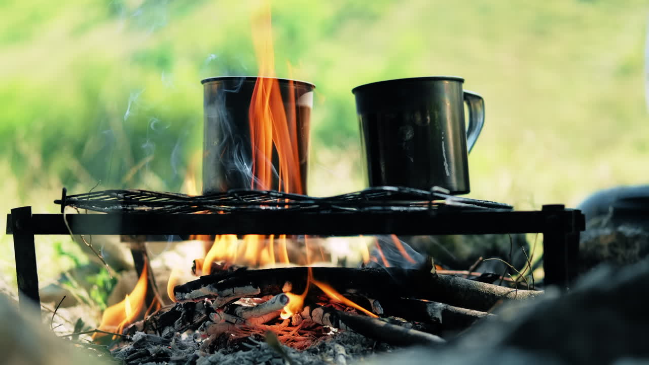 Water in two tin mugs is boiling on a grill grid above the campfire