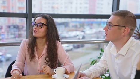 young people wearing glasses sit at table and speak looking up at standing person. teamwork in the office at the backdrop of panoramic windows and cityscape.