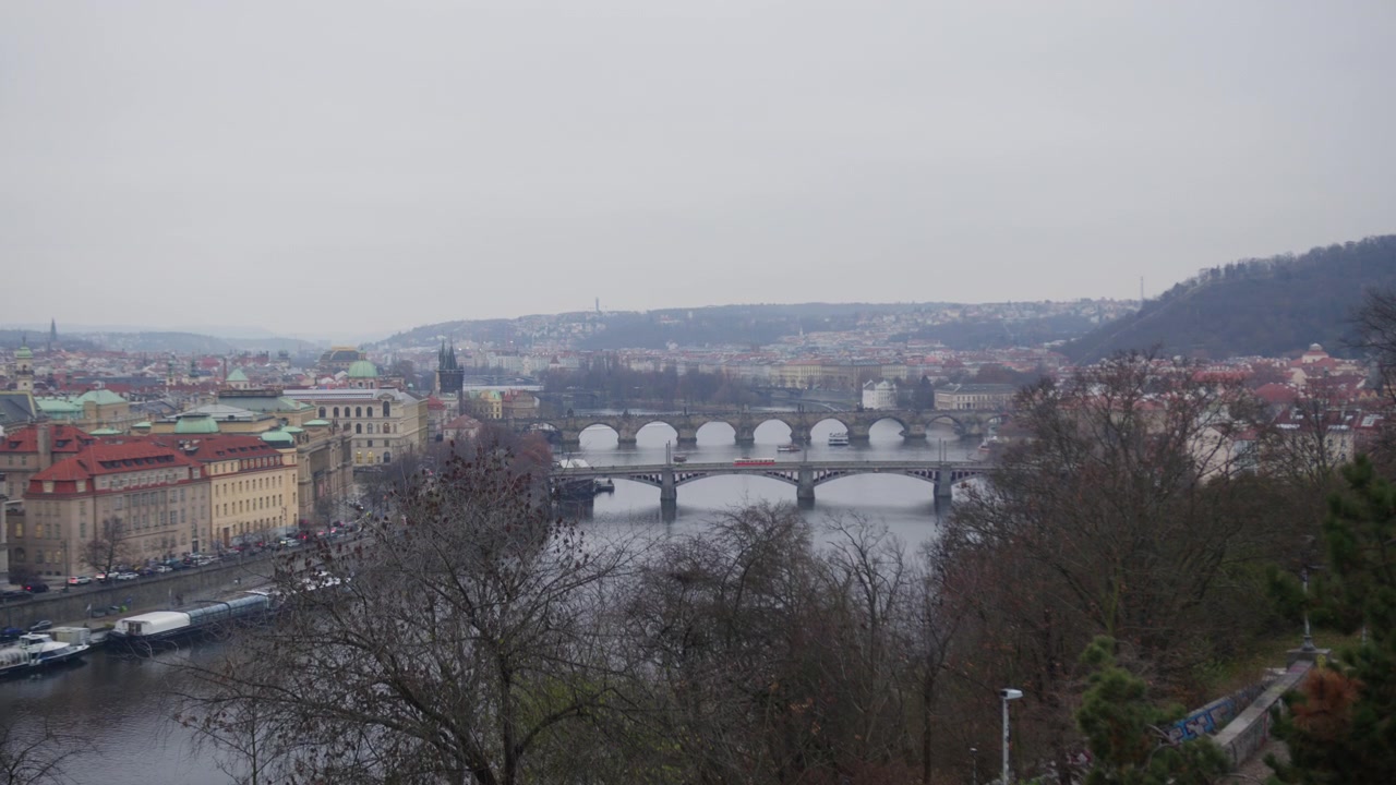 Panorama elevato sulla Moldava dove il Ponte Carlo e una serie di altri ponti attraversano l'acqua, con i tetti rossi e le cupole verdi della Città Vecchia che sfumano nella foschia invernale.