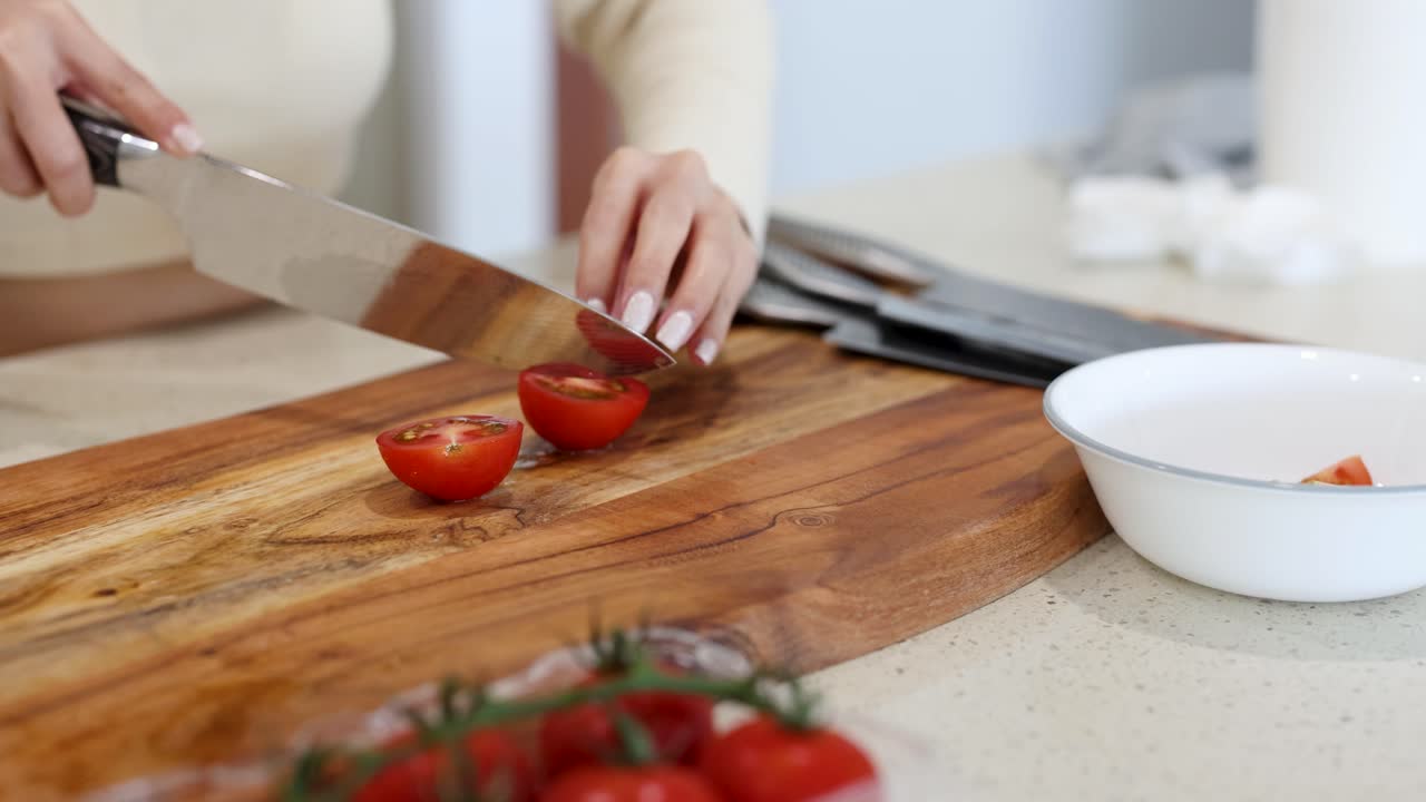 A person slices tomatoes on a wooden board in a bright kitchen, placing them into a white bowl