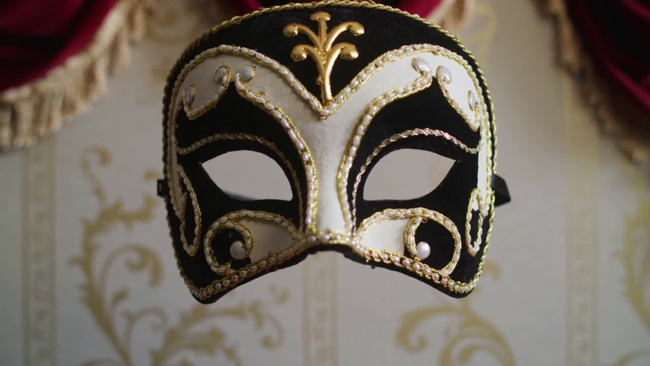 La telecamera ingrandisce, una maschera si avvicina su carta da parati con motivi a vite dorati e un drappo rosso, mettendo in risalto le perle