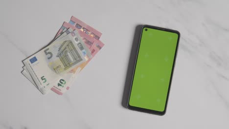 Tiro-De-Moneda-Por-Encima-De-La-Cabeza-Persona-Poniendo-Billetes-Y-Monedas-En-Euros-Junto-A-Un-Teléfono-Móvil-Con-Pantalla-Verde