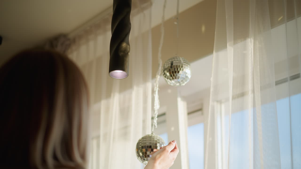 Woman decorating interior by hanging shiny disco balls near window with sheer curtains, natural daylight illuminating reflective surfaces while spotlight above enhances sparkling festive atmosphere at home