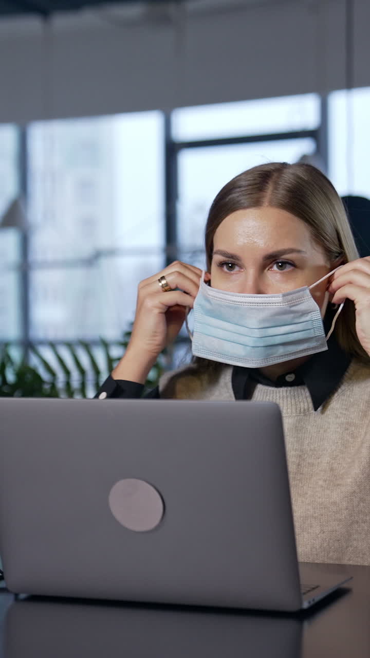 Girl in office puts a mask on and fits it to her face. Long-haired female employee wears mask and continues her work at the computer. Vertical video