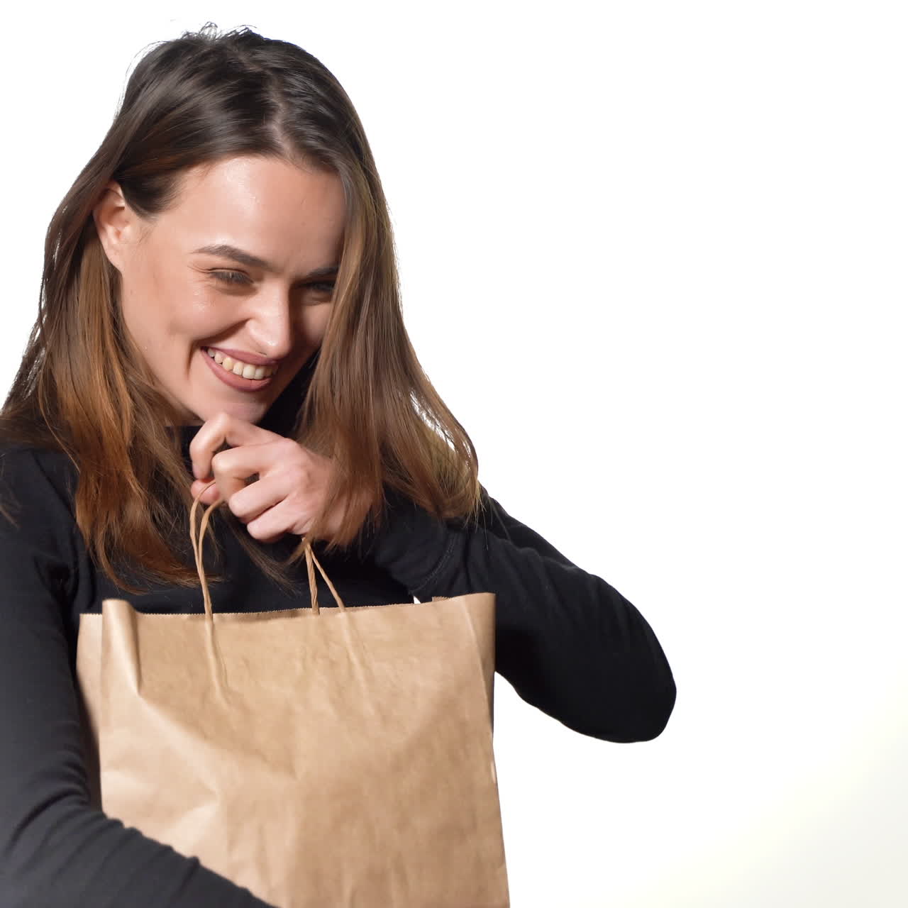 Happy woman with long hair is holding a paper bag with gift in her hands and looking inside of it on white background. The girl is admiring what she see.