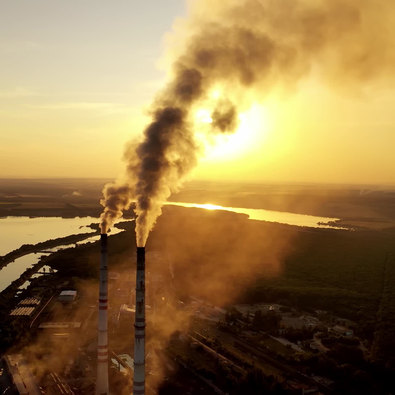 Dirty emissions coming out of industrial pipes. Factory surrounded by nature in the evening. Thick smoke going into th air from manufacturing at sunset. Pollution.