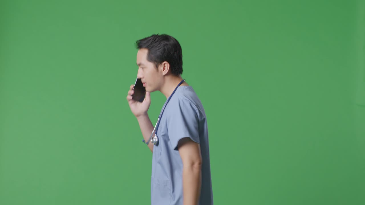 병원 에서 녹색 스크린 배경 에 걸어 다니는 동안 스테토스코프 를 들고 스마트폰 으로 이야기 하는 아시아인 남성 의사 의 측면 시각