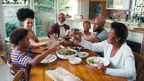 Multi-Generation-Family-Sitting-Around-Table-Doing-Cheers-With-Water-Before-Meal-At-Home-Together