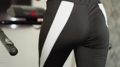 mujer hace ejercicios en la máquina de entrenamiento en casa de cerca. dama elegante en pantalones deportivos corre rápido en la cinta de correr para fortalecer los músculos glúteos en el gimnasio doméstico. cuidado del cuerpo