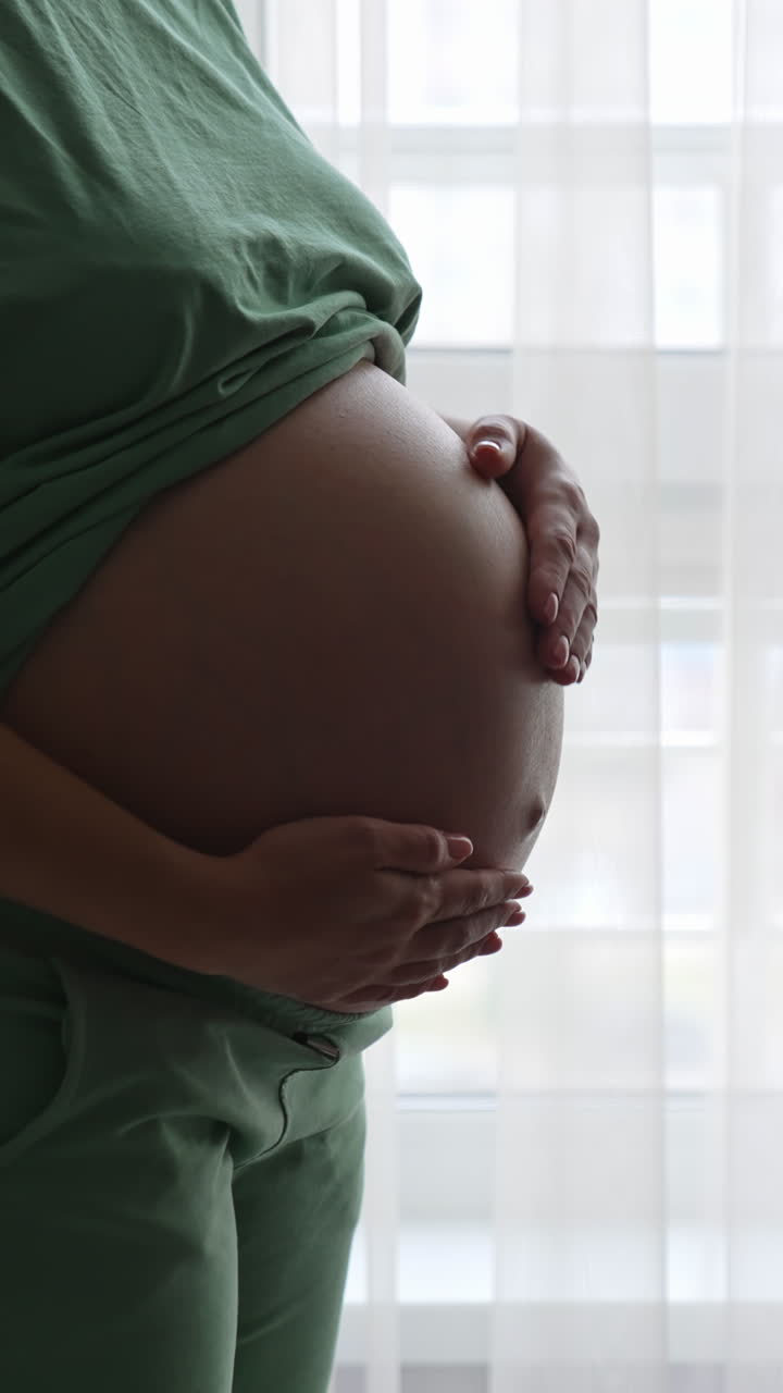 Future mom standing at the window and touching her belly gently. Expecting woman in comfortable clothes strokes her tummy lovingly. Vertical video