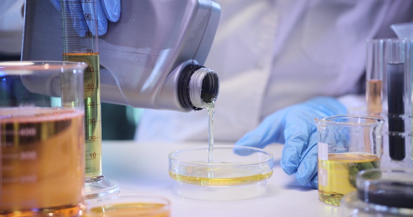 Lab technician pours yellow engine oil into glass dish
