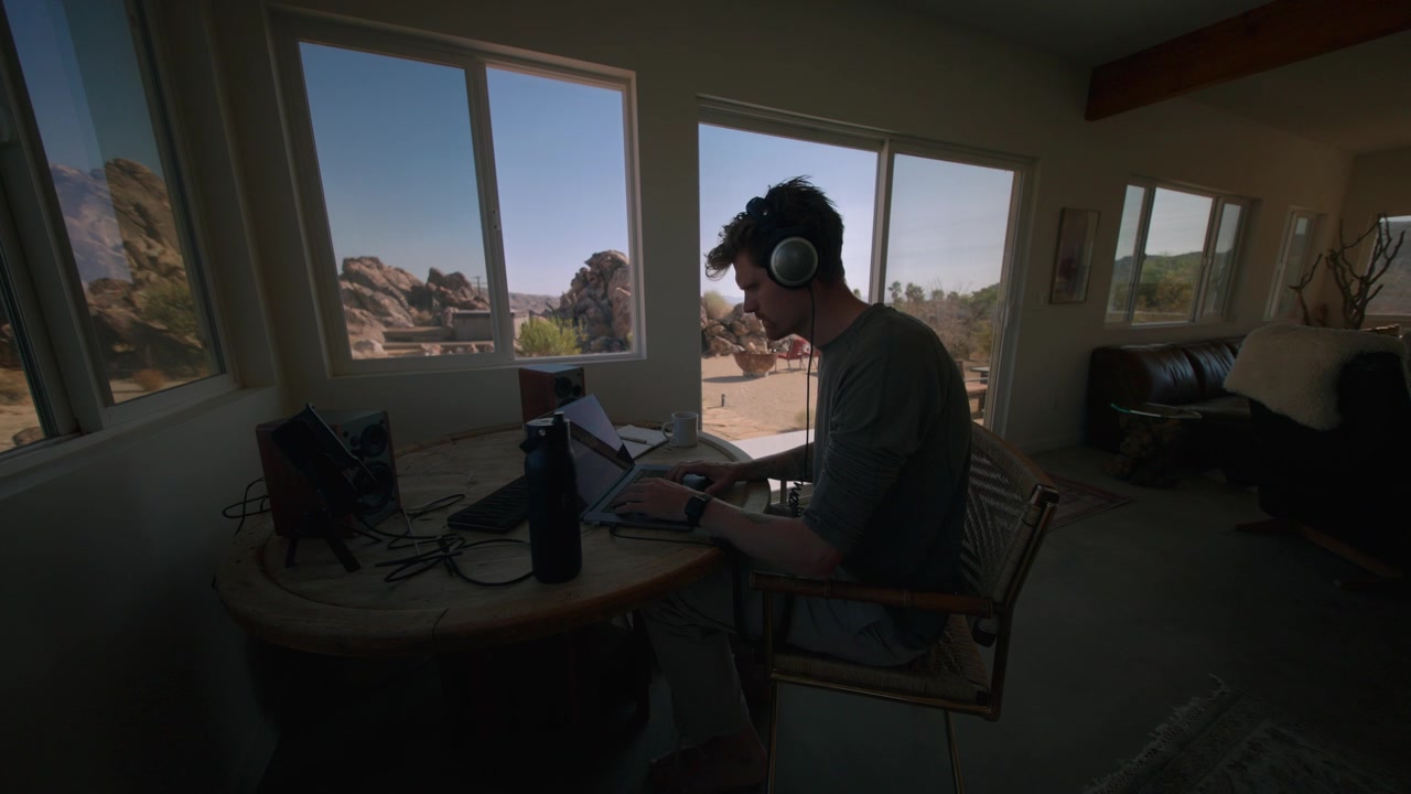 Indossando le cuffie, un uomo regola l'attrezzatura audio a un tavolo con vista sul deserto dalla finestra.