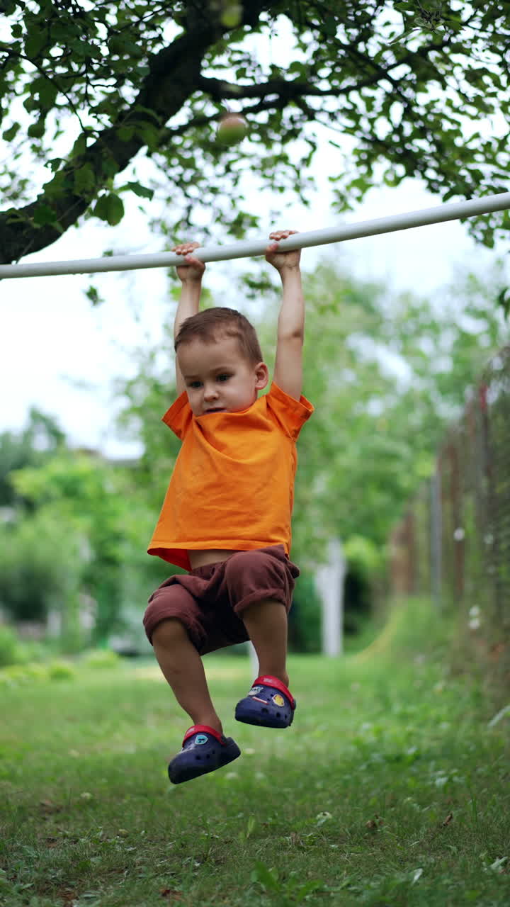 Little baby boy exercising on the horizontal bar outdoors. Caucasian kid doing sport. Vertical video.