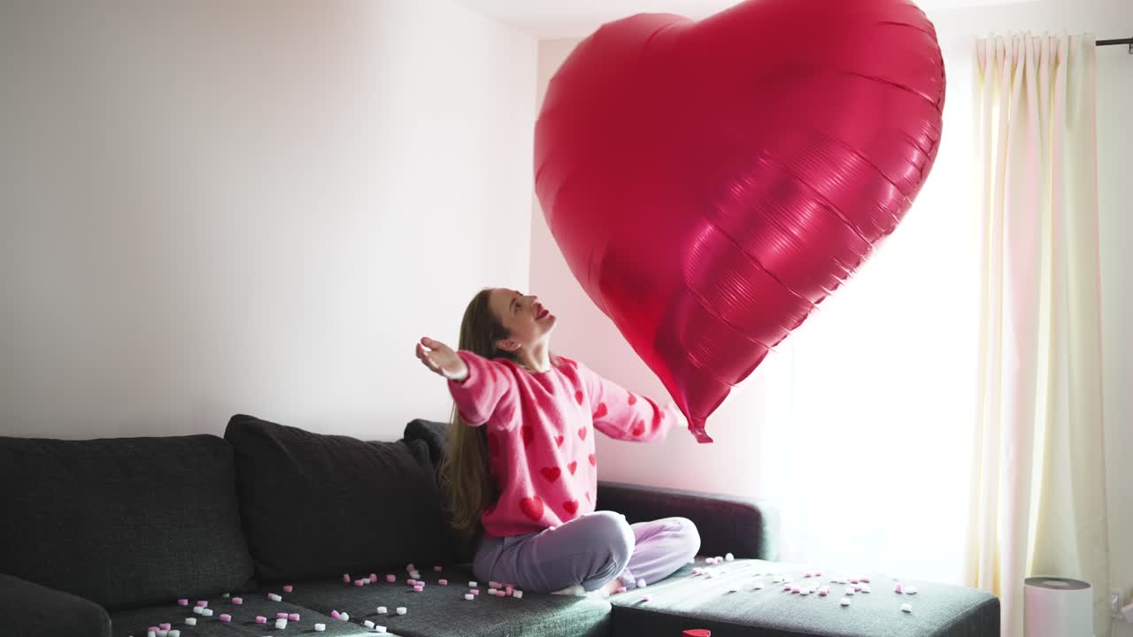 La giovane donna si rilassa nel suo salotto, sorridendo e godendosi San Valentino con un grande palloncino a forma di cuore sul divano, circondata da un'illuminazione tenue e un semplice arredamento romantico