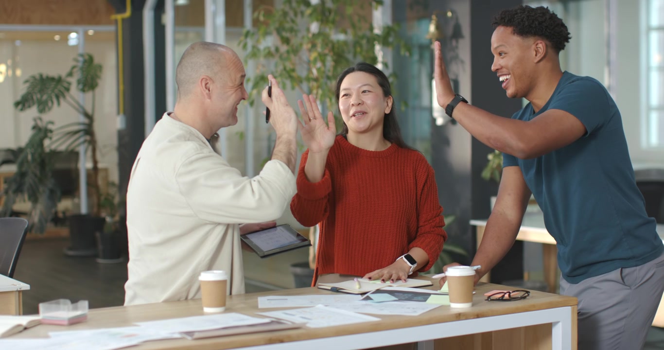 Diverse coworkers confirming layout at high table in office, annotating on tablet, then high-fiving
