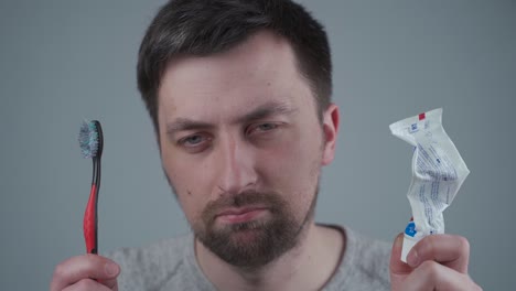 man is upset about not buying oral hygiene products on time and holding them in his hands empty tooth paste and used tooth brush in studio on gray background. means for cleaning teeth and mouth