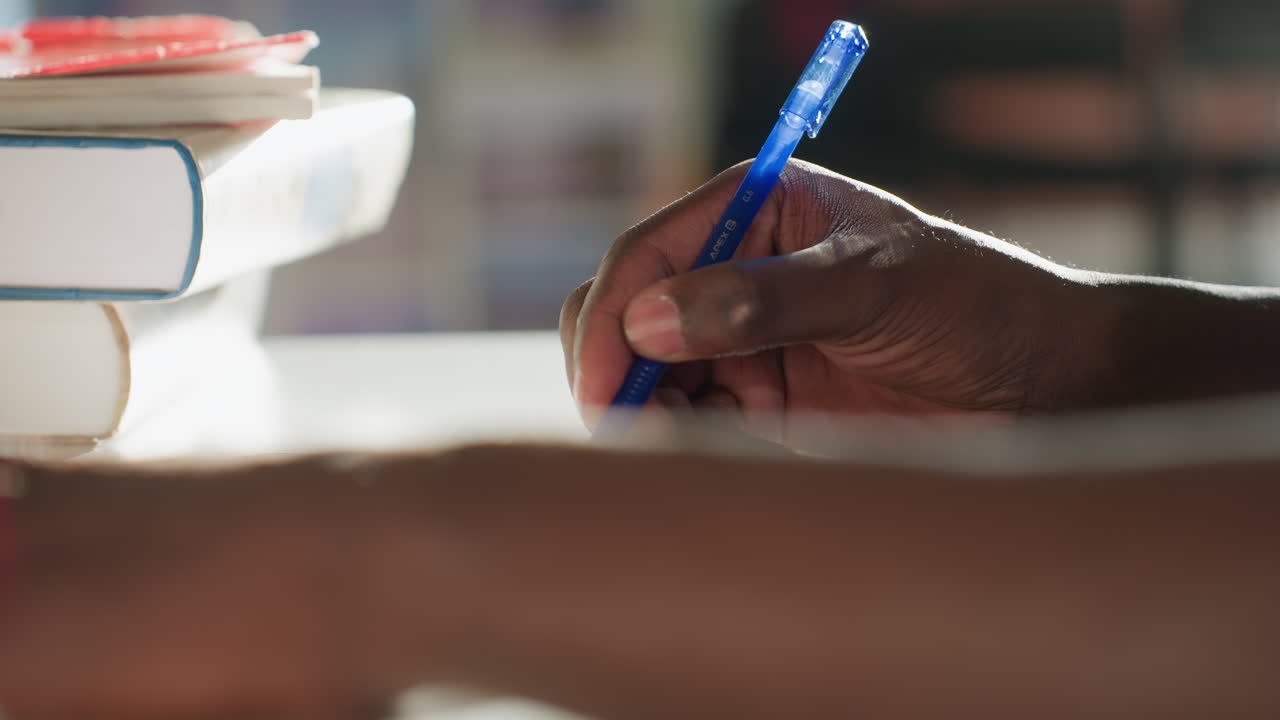 Close up of dark skinned hand holding blue pen while writing beside stacked books, blurred background with warm sunlight highlighting scene, capturing focused moment of note taking and study