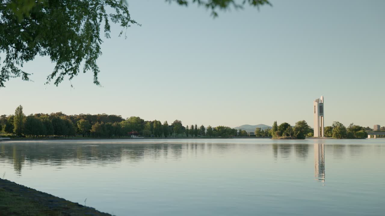 A closer view of the National Carillon rising over Lake Burley Griffin, its structure illuminated by the morning sun, with surrounding trees and calm waters adding to the tranquil Canberra scene.