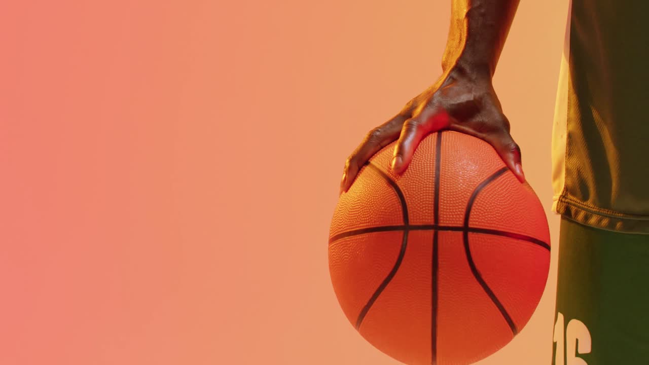 el video de la sección media del jugador de baloncesto afroamericano sosteniendo la pelota sobre un fondo naranja.