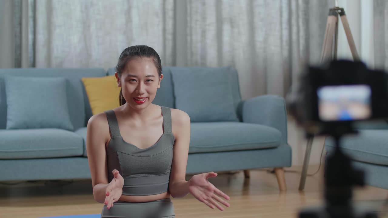 joven entrenadora asiática mujer en ropa deportiva agitando la mano y hablando a la cámara mientras graba ejercicio de enseñanza en casa