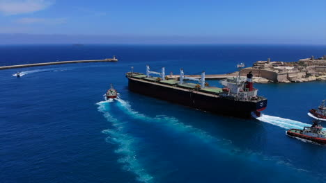 Unique-aerial-shot-of-a-giant-cargo-ship-leaving-the-harbour-of-Valetta,-Malta-with-the-help-of-three-tugboats