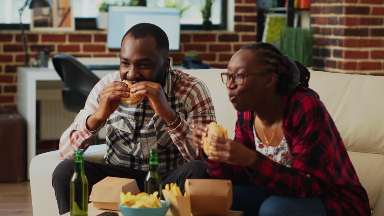 Modern young couple eating burgers together on couch
